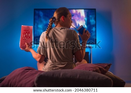 A blonde girl in pajamas watches TV at night. Insomnia. She is holding a pack of popcorn and a carbonated drink with a straw. Neon light. Watching TV shows and series, quarantine. Royalty-Free Stock Photo #2046380711