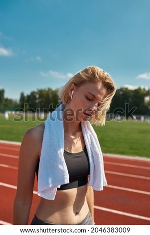 Young sportswoman with towel around her neck listening to music using earbuds while standing on the track at stadium on a sunny day. Outdoor workout concept