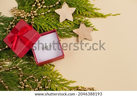 a white gold ring with a large gemstone in a red gift box with a bow on the background of a green thuja branch with a golden garland and wooden decor on a blurred background with a place for text Royalty-Free Stock Photo #2046388193