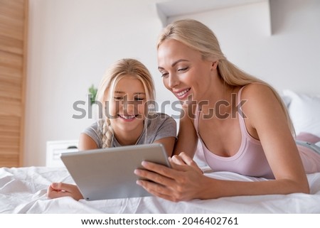 Beautiful young happy woman mother babysitter nanny tutor and her little daughter using digital tablet for watching cartoons e-learning while lying in bed at home