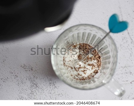 Before pouring hot boiled water from black kettle into a glass mug with three in one instant coffee mix and heart shaped teaspoon on white cement kitchen countertop background. (selective focus)