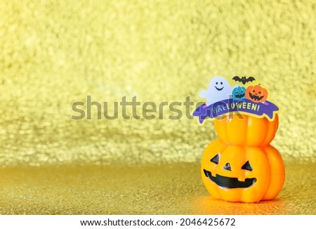 Studio photography depicting a smiling halloween jack o lantern pumpkin head with cardboard cutout of a funny ghost and bats with the word Halloween! against a glitter golden background.