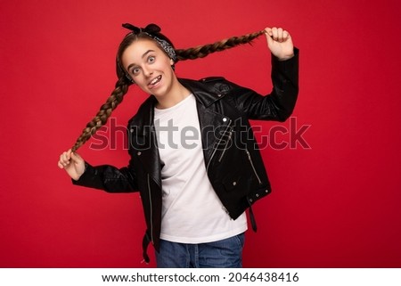 Photo shot of beautiful happy smiling brunette little girl with pigtails wearing trendy black leather jacket and white t-shirt for mockup standing isolated over red background wall looking at camera