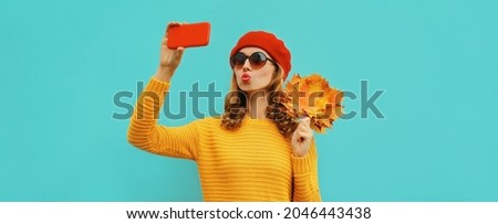 Autumn portrait of beautiful young woman taking selfie by smartphone with yellow maple leaves wearing a sweater, red french beret on blue background