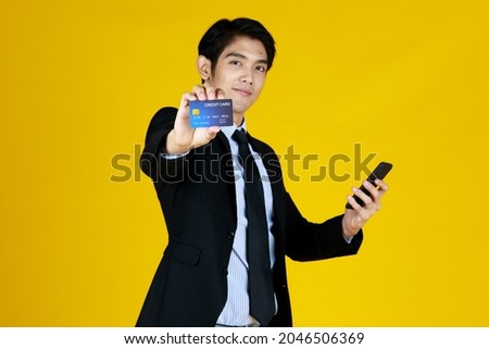 Young man in black formal suit. Stand and show credit card with confidence gesture. While other hand holds mobile phone. Yellow background make photo look attractive