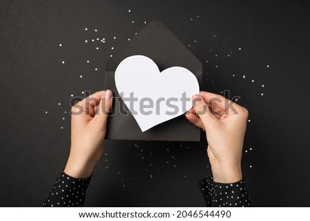 First person top view photo of girl's hands in polka dot shirt holding black open envelope with white paper heart over sequins on isolated black background with blank space