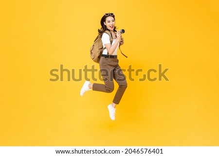 Young pretty Asian woman tourist backpacker smiling and jumping with camera in hand isolated on yellow studio background