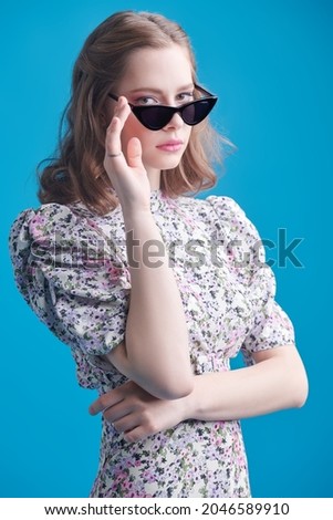 Beautiful girl with romantic wavy hair, soft pink make-up in a light summer dress and sunglasses looking at the camera. Studio portrait in a blue background. Summer beauty and fashion. Happy people.