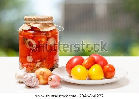 Tomatoes preserved in glass jars, fresh tomatoes on a plate, onions and garlic. Pickled tomatoes on the blurred natural background. Homemade preserves.