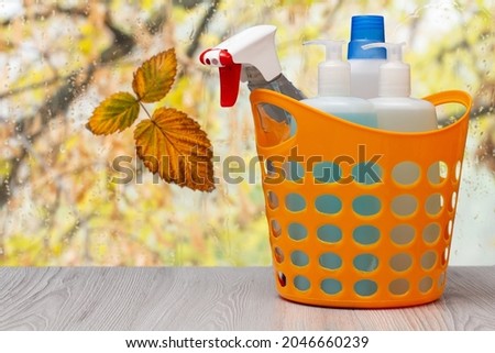 Plastic basket with bottles of dishwashing liquid, glass and tile cleaner, detergent for stoves in front of the window with water drops and autumn leaves. Washing and cleaning concept.