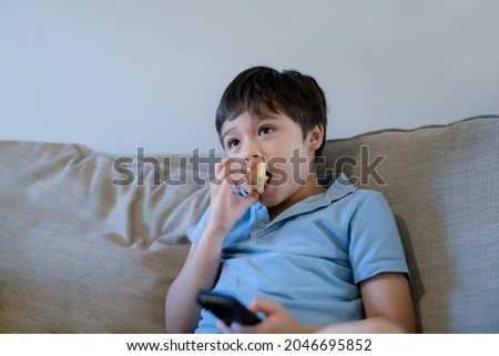 Happy boy bitting red apple while watching cartoon on TV, Mixed race school kid relaxing at home after back from school, Positive child sitting on sofa eating fruit for snack in living room.