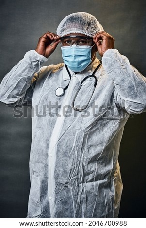 Young African male doctor putting on a blue surgical face mask with stethoscope around his neck wearing a disposable white lab coat and hairnet in studio