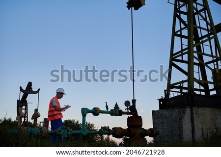 Side view of oil well worker who writing results of pipeline inspection on clipboard. Engineer making notes while servicing the activities of oil rig against blue sky.