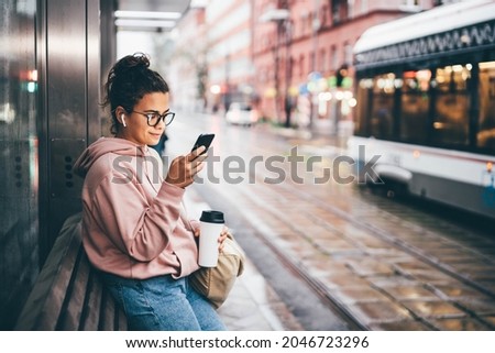 Young woman with glasses and mobile phone waits for bus and gets into salon on public transport stop against large city blurry street in rainy evening side view Royalty-Free Stock Photo #2046723296