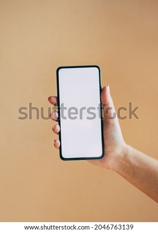 An Anonymous Woman Holding Mobile Phone with Empty Screen on Brown Background.  
Close up photo of female hand showing smartphone with blank screen isolated over a beige background.