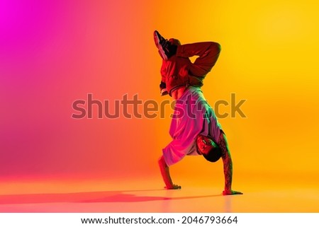Headstand. Portrait of young stylish man, break dancing dancer training in casual clothes isolated over gradient pink yellow background. Youth culture, movement, street style and fashion, action. Royalty-Free Stock Photo #2046793664