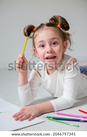 funny little girl with glasses and an unusual hairstyle is lying on the wooden floor and drawing in an album with colored pencils. home education. kindergarten. space for text