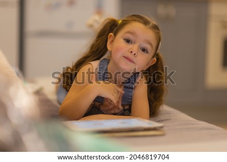 Smiling little girl lying on comfortable couch, enjoying playing online game on digital tablet computer. Addicted to technology, happy small kid using funny applications, web surfing information.