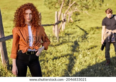 Portrait of cute curly redhead lady posing in green field near forest, at sunny day in rural environment. handsome guy in the background, spending summer vacations together. travel concept