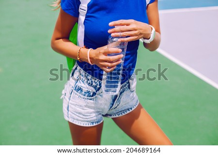 Young healthy sportive tan girl in jeans shores and navy t-shirt, holding bottle of water in her hands, sports ground background.