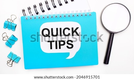 Office table with blue notepad, paper clips, magnifying glass and white card with text QUICK TIPS. Top view, flat lay. Stylish workplace