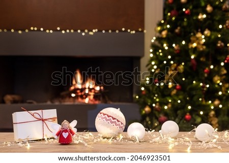 Cozy Christmas atmosphere with angel, decorations and gifts on wooden table. In the background blurred the fireplace with fire, the Christmas tree and Christmas decorations. Selective focus