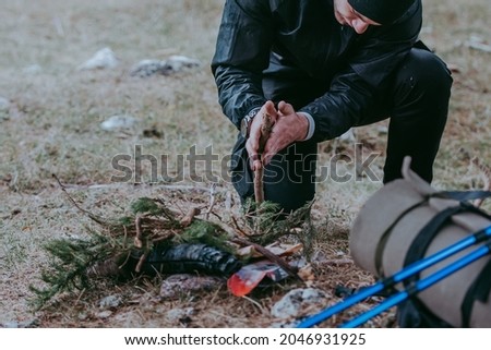 Serious young bearded hiker in cap setting fire in the forest using a wood sticks. Various methods of starting a campfire. Survival skills. Royalty-Free Stock Photo #2046931925