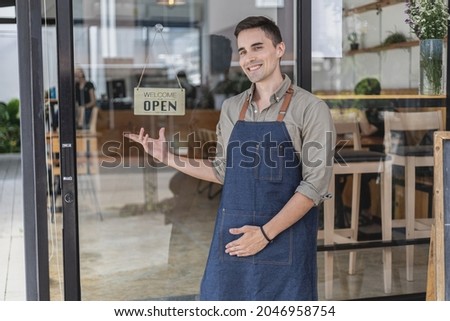 A male cafe employee standing in front of the store greets customers and has a sign that says open to show that the shop is open, a male employee opens the shop to serve food and beverages.