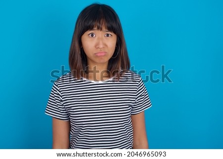 Displeased Young beautiful asian girl wearing striped t-shirt over blue background frowns face feels unhappy has some problems. Negative emotions and feelings concept