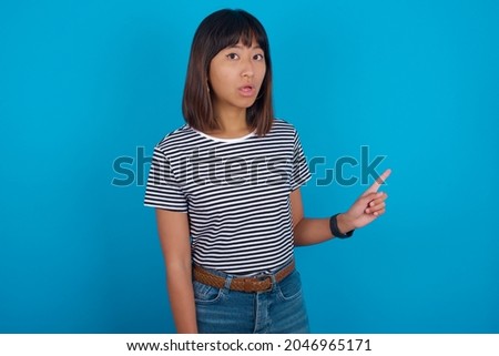 Emotive Young beautiful asian girl wearing striped t-shirt over blue background keeps jaw dropped from shock demonstrates amazing promo points right on blank space demonstrates big shopping sale.