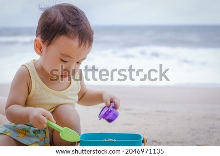 Asian boy child wear yellow vest playing sand with his bucket while sitting on the beach in front of sea. Phuket, Thailand. Family Holiday, summer trip concept. Royalty-Free Stock Photo #2046971135