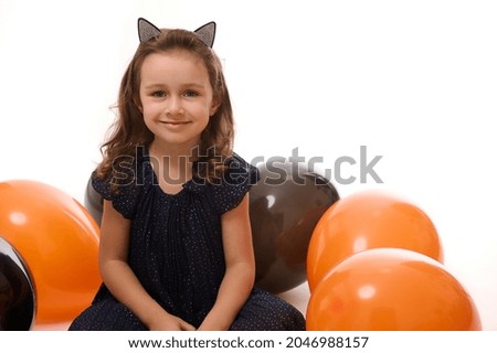 Cheerful winsome beautiful little girl in black carnival dress and hoop with cat ears sitting on a white surface with bi colored balloons, smiles looking at camera. Halloween decoration. Copy space