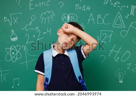 Young sick ill male kid school boy 5-6 years old in t-shirt backpack put hand on forehead isolated on green wall chalk blackboard background studio. Childhood children kids education lifestyle concept