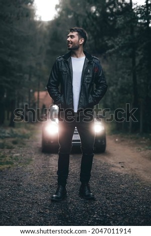 Portrait of white male person wearing leather jacket looking to the side in the woods and smiling while standing in front of his car with lights on.