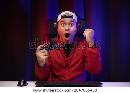 Excited and shocked face of Asian gamer with headphone holding joystick playing video game online sitting on chair at living room. Indian professional gamer streaming on social playing game very fun Royalty-Free Stock Photo #2047055438