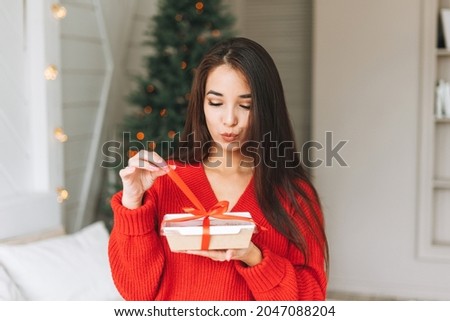 Young beautiful asian woman with dark long hair in cozy red knitted sweater and santa hat with present gift box with red ribbon sitting on bed in room with Christmas tree