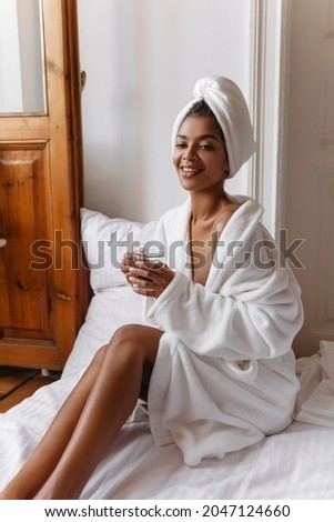  dark-skinned lady without makeup with towel on her head holds glass cup of coffee. woman in white robe posing in bedroom while sitting on made bed. Royalty-Free Stock Photo #2047124660