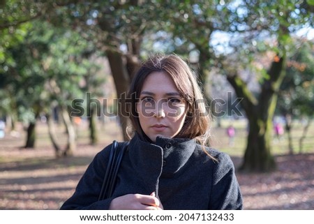 Young woman in a coat in a park. Untouched photo with light makeup, close up direct eye contact with the camera with a neutral expression. Dynamic photo with blurred nature background.