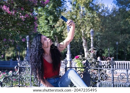 Middle-aged adult Hispanic woman with black curly hair taking a photo with her cell phone. Technology and selfie concept.