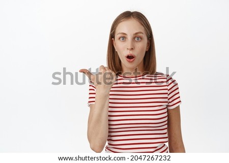 Wow look that way. Impressed blond woman in awe, gasp and stare at camera speechless, pointing thumb left, showing sale advertisement, standing in striped t-shirt over white background