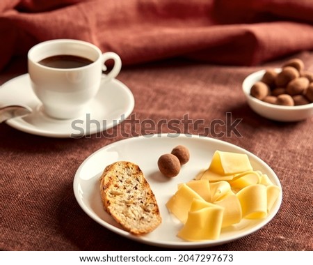 Sliced cheese, toast and sweets lie on a white plate. The plate is on a brown tablecloth. Behind the plate is a cup of coffee and a small bowl of sweets. In the background is a terracotta napkin