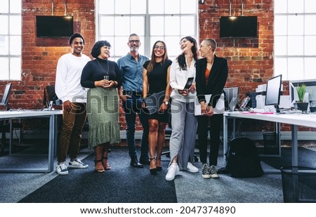 Happy group of businesspeople laughing cheerfully in a modern workplace. Diverse group of colleagues enjoying working together in an office. Successful businesspeople standing together. Royalty-Free Stock Photo #2047374890