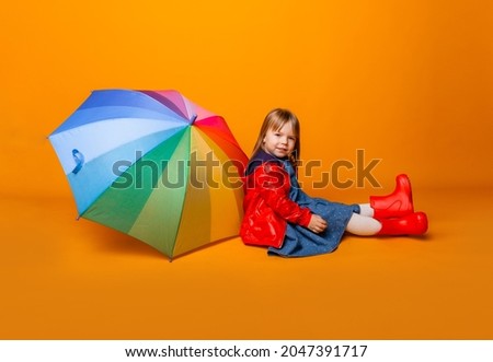 A smiling child girl in a red jacket and boots, holding an umbrella, stands on a yellow background. little girl in bright clothes holds a rainbow umbrella.