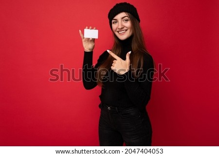 Photo of beautiful young brunette woman wearing black sweater and hat isolated on red background holding credit card looking at camera and pointing finger at plasctic card
