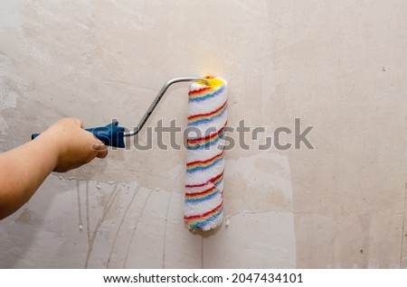 A woman holds a paint roller and impregnates the walls of the room with a primer. Preparing the walls for painting. Inside the room. Selective Focus. Royalty-Free Stock Photo #2047434101