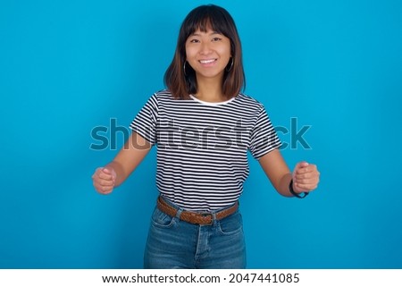 Young beautiful asian girl wearing striped t-shirt over blue background makes phone gesture, says call me back again, has glad expression.