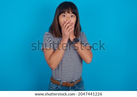 Shocked Young beautiful asian girl wearing striped t-shirt over blue background points at you with stunned expression