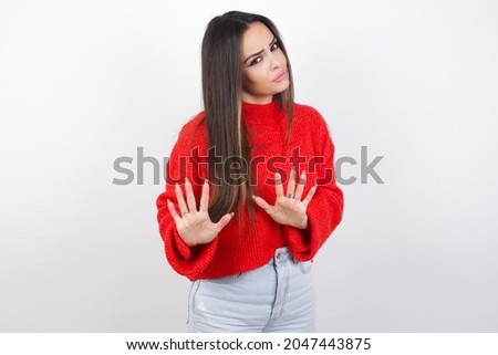 Afraid young beautiful brunette wearing red knitted sweater over a white wall, makes terrified expression and stop gesture with both hands saying: Stay there. Panic concept.