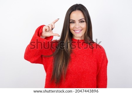 young beautiful brunette wearing red knitted sweater over a white wall smiling and gesturing with hand small size, measure symbol.