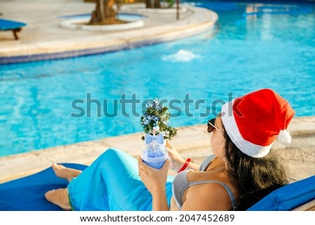 A woman in a Santa hat and sunglasses and a swimsuit in a sun lounger by the pool does not see her face. Horizontal photo